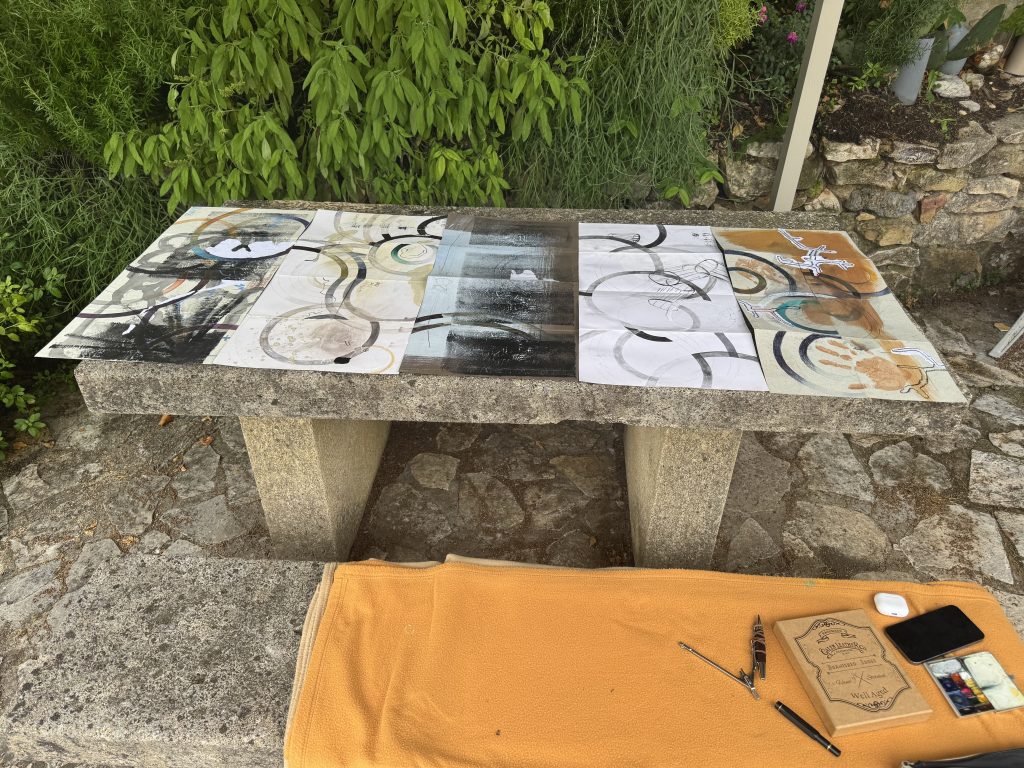 Artist's workspace showing multiple accordion-fold artworks in progress with watercolor palette and brushes outdoors