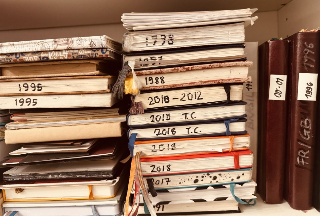 Stefani Peter's stack of personal journals and sketchbooks dated from 1988 to 2018, showing three decades of daily practice and writing, with colored ribbon bookmarks