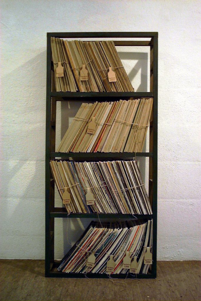 Studio documentation photograph of custom black bookshelf (Buchregal) displaying Stefani Peter's complete Books of Days collection created between 1992-2000, totaling approximately 350 accordion-fold books. The books are stored in their compact folded state, organized chronologically on four shelves and bundled with string, with hand-labeled tags marking years: 1992, 1993, 1994, 1995, 1996, 1997, 1998, 1999, and 2000. Each shelf shows the accumulation of roughly 8-9 years of daily practice, with visible color variations in the book edges revealing the diverse palettes used across different periods. Photographed in the artist's studio, Fürth, Germany, 2001—shortly before immigration to Canada.